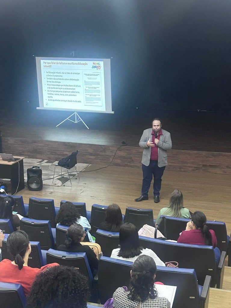 Renan Sargiani em palco de auditório, com plateia sentada e slide sobre leitura e escrita na Educação Infantil