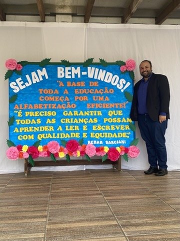 Renan Sargiani ao lado de um painel de boas-vindas decorado com flores de papel, em sala de formação
